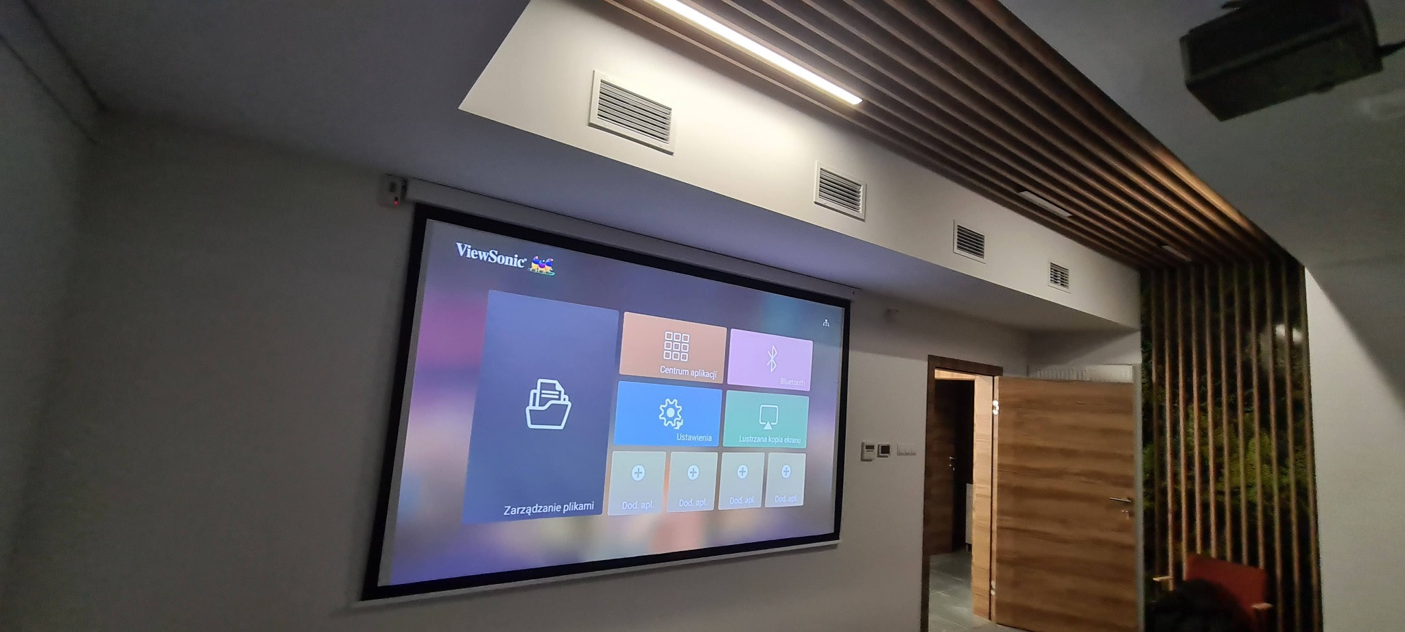 Sala konferencyjna z zainstalowanym projektorem ViewSonic i ekranem projekcyjnym wyświetlającym menu aplikacji, widoczne kratki wentylacyjne w suficie i dekoracyjna ściana z drewnianych listew...