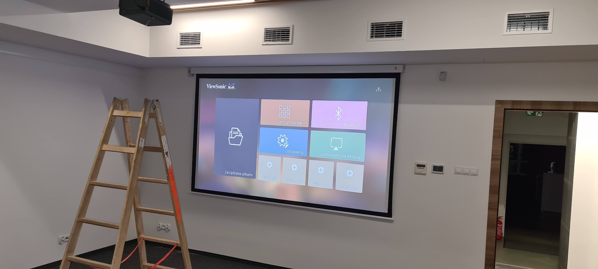 Sala konferencyjna z rozwijanym ekranem projekcyjnym ViewSonic, na którym wyświetlany jest interfejs użytkownika; obok stoi drewniana drabina, a w tle widoczne kratki wentylacyjne.