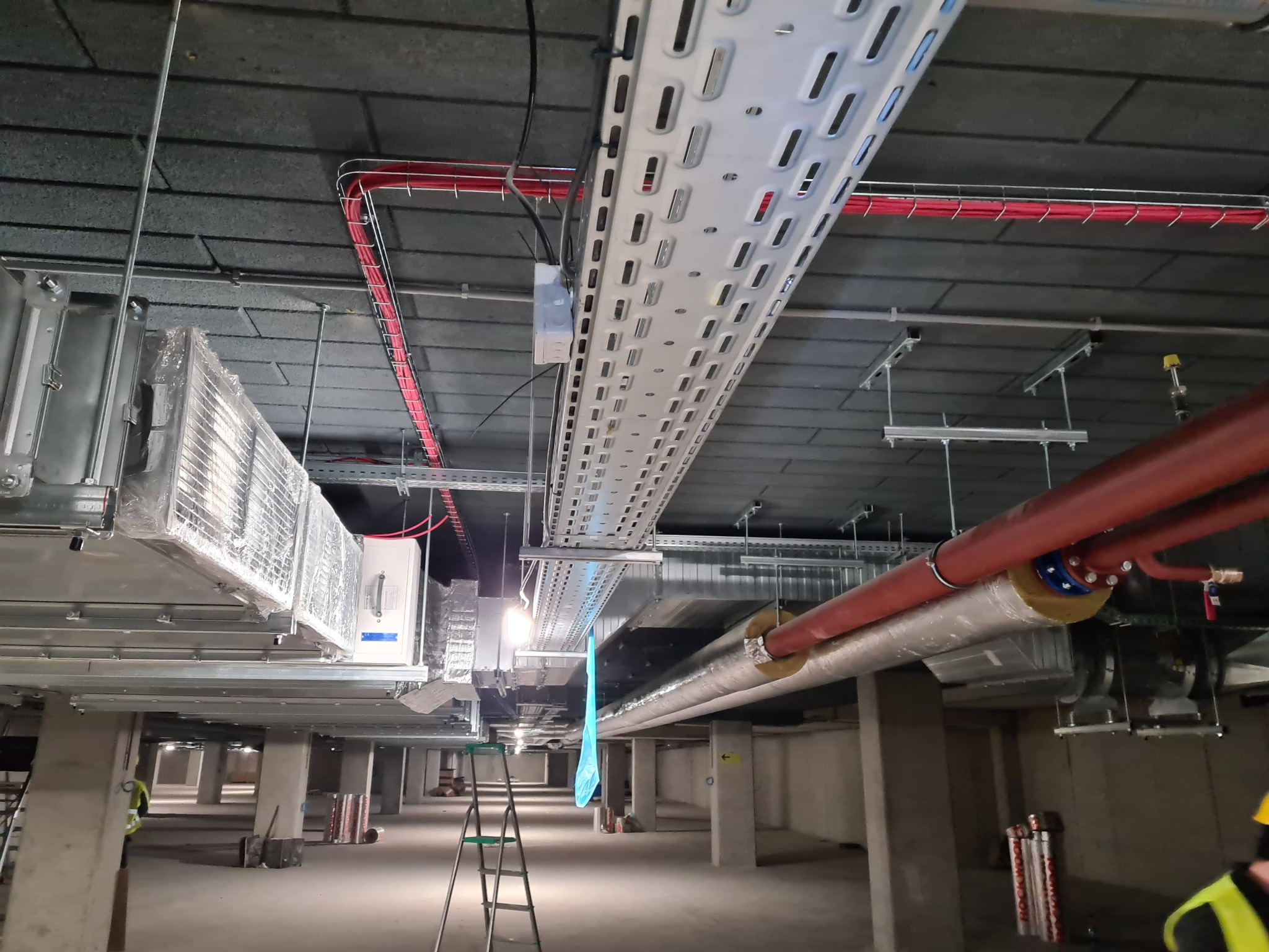 Rozbudowana instalacja w hali: koryta kablowe z przewodami, rury izolowane termicznie, elementy wentylacyjne i drabina monterska na tle surowego betonu.