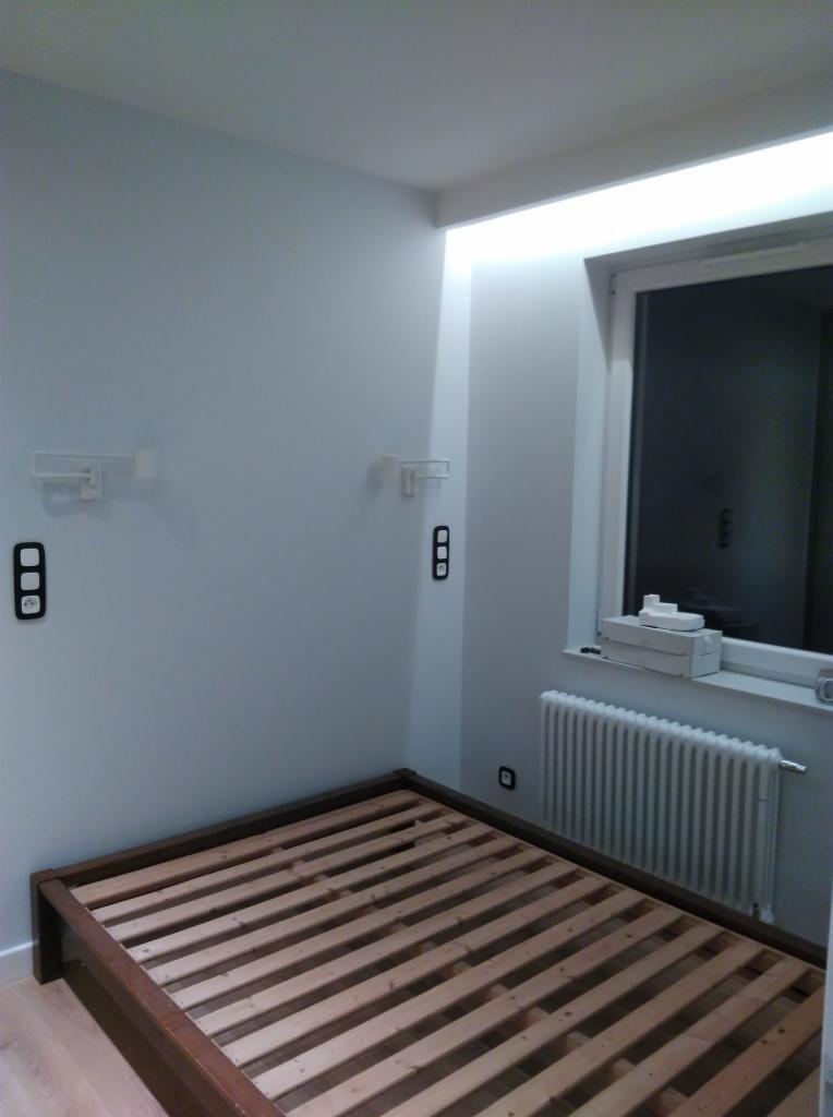 Pusty, drewniany stelaż łóżka w pokoju z białymi ścianami, grzejnikiem pod oknem i przygotowanymi punktami oświetleniowymi na ścianie.