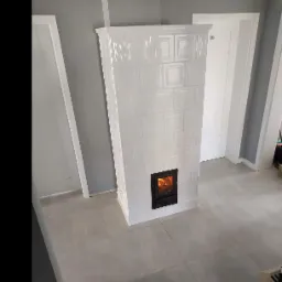 Biały piec kaflowy z paleniskiem, płomienie widoczne przez czarne drzwiczki, na tle szarej ściany i podłogi z płytek.