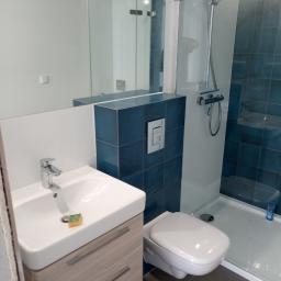 Glazura, terakota i inne - Nowoczesna łazienka po remoncie: biała umywalka z szafką w kolorze jasnego drewna, toaleta podwieszana, kabina prysznicowa z niebieskimi płytkami, szara podłoga i mała gąbka na umywalce.