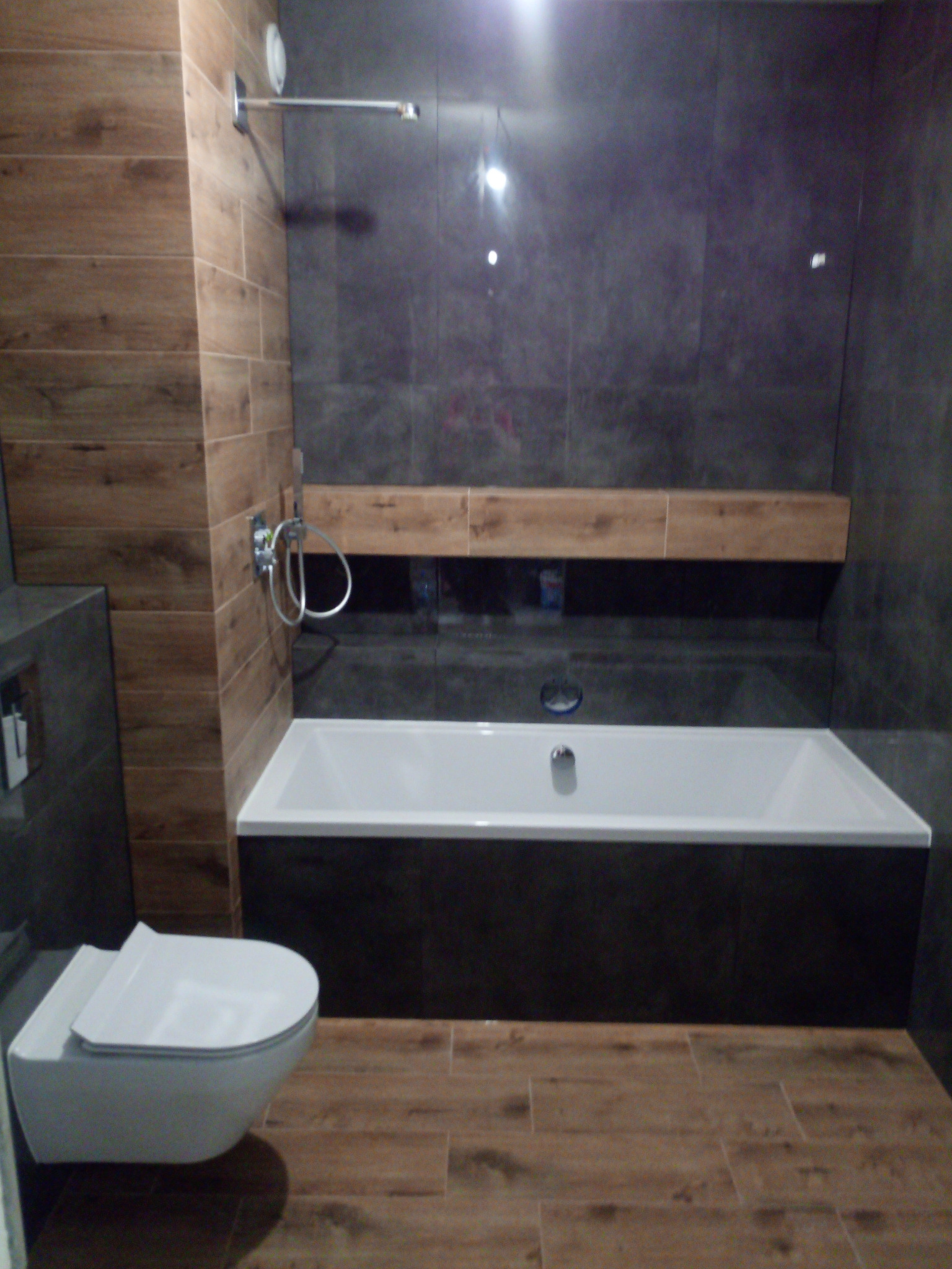 Nowoczesna łazienka z drewnopodobnymi płytkami na podłodze i ścianie, grafitowymi płytkami na ścianie z wanną, wisząca toaleta z deską, bateria prysznicowa z deszczownicą, półka nad wanną.