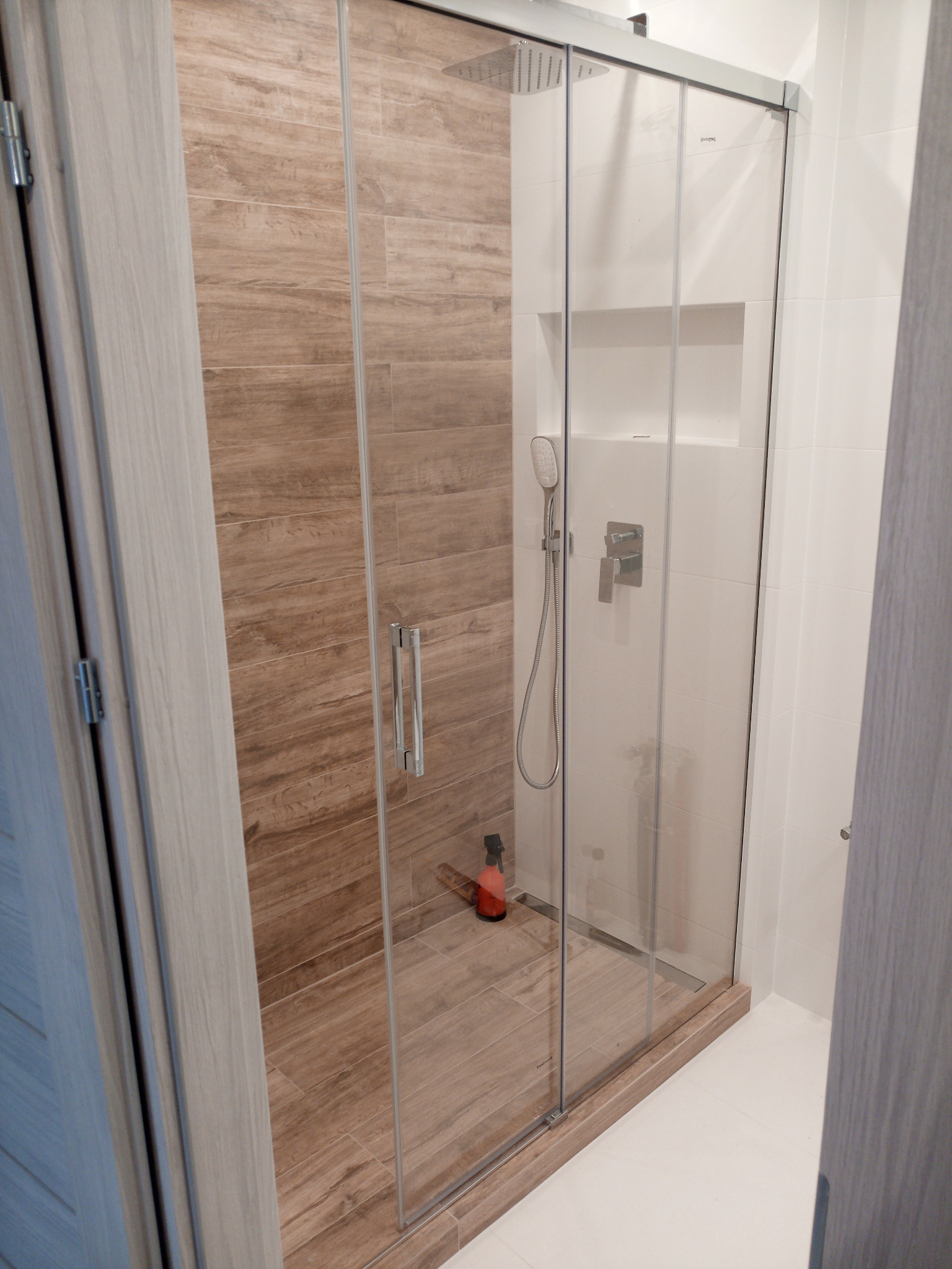 Nowoczesna kabina prysznicowa z przesuwnymi drzwiami ze szkła hartowanego, wykończona płytkami imitującymi drewno i białymi płytkami, z widocznym dozownikiem na płyn do mycia pod prysznicem.
