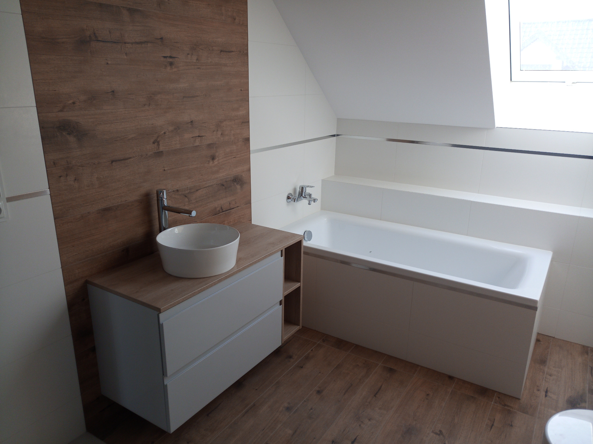 Nowoczesna łazienka na poddaszu: biała wanna obudowana płytkami, umywalka nablatowa na szafce z drewnianym blatem, ściana z paneli imitujących drewno, podłoga z płytek drewnopodobnych, okno dachowe.