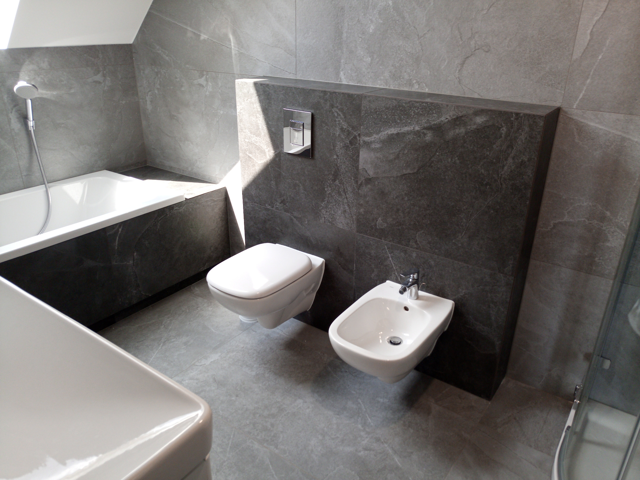 Nowoczesna łazienka w odcieniach szarości z białą armaturą: wisząca toaleta, bidet i wanna z prysznicem. Na ścianach i podłodze płytki imitujące kamień. Widoczny panel z przyciskiem spłukiwania...