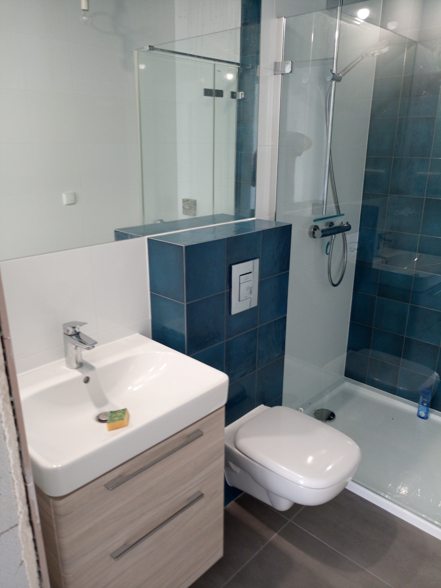 Nowoczesna łazienka po remoncie: biała umywalka z szafką w kolorze jasnego drewna, toaleta podwieszana, kabina prysznicowa z niebieskimi płytkami, szara podłoga i mała gąbka na umywalce.