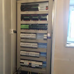 Otwarta szafa sterownicza z licznymi modułami, bezpiecznikami i przewodami, zasilana bateryjnie; widoczny napis LOXONE na niektórych modułach i naklejka ostrzegawcza PV.