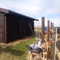 Usługi Wielobranżowe Agata Opioła - Budowa wiaty drewnianej z ciemnego drewna i słupów, widoczne narzędzia i drabina, pracownik w oddali.