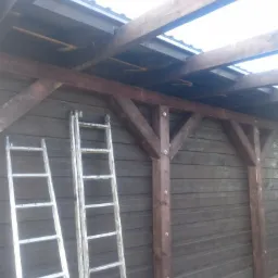 Wznoszona drewniana konstrukcja wiaty garażowej z brązowymi belkami i przezroczystym zadaszeniem, widoczne dwie aluminiowe drabiny opierające się o ciemną, drewnianą ścianę. Etap budowy.