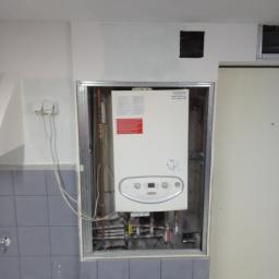 Usługi Wielobranżowe Agata Opioła - Biały piec gazowy zamontowany w metalowej ramie, z widocznymi rurami i instalacją, na tle szarych płytek i białej ściany z otworem wentylacyjnym.