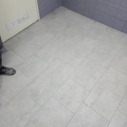 Usługi Wielobranżowe Agata Opioła - Ułożone jasnoszare kwadratowe płytki podłogowe w rogu pomieszczenia, z fragmentem ściany wyłożonej szarymi płytkami i częścią białych drzwi widoczną po lewej stronie, oraz butem roboczym w lewym...