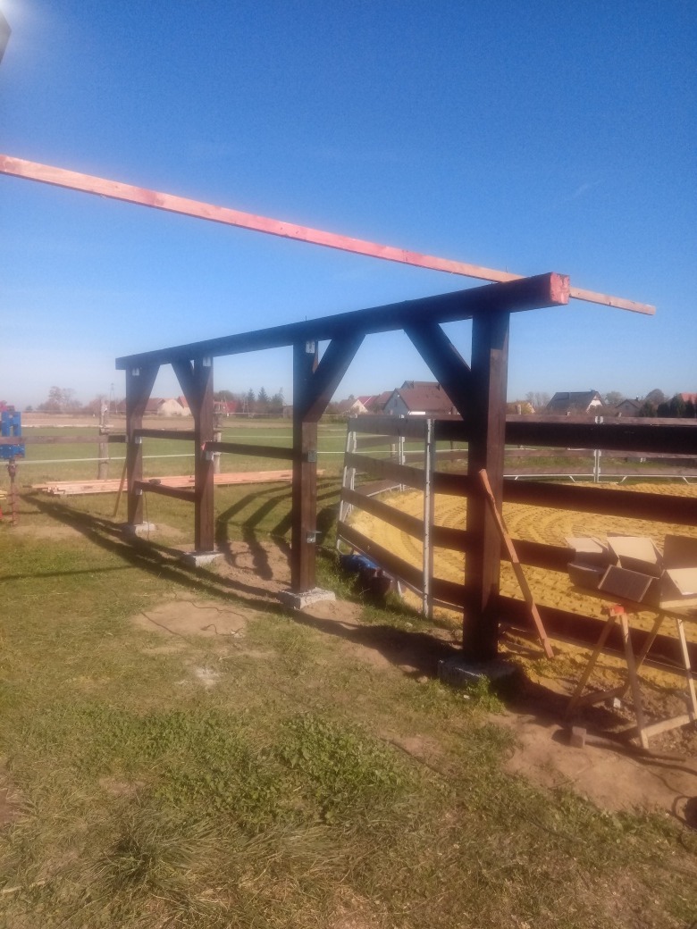 Wznoszona konstrukcja drewniana z widocznymi słupami na betonowych fundamentach i niezabezpieczonymi deskami na górze, sugerująca budowę zadaszenia lub wiaty na zewnątrz.