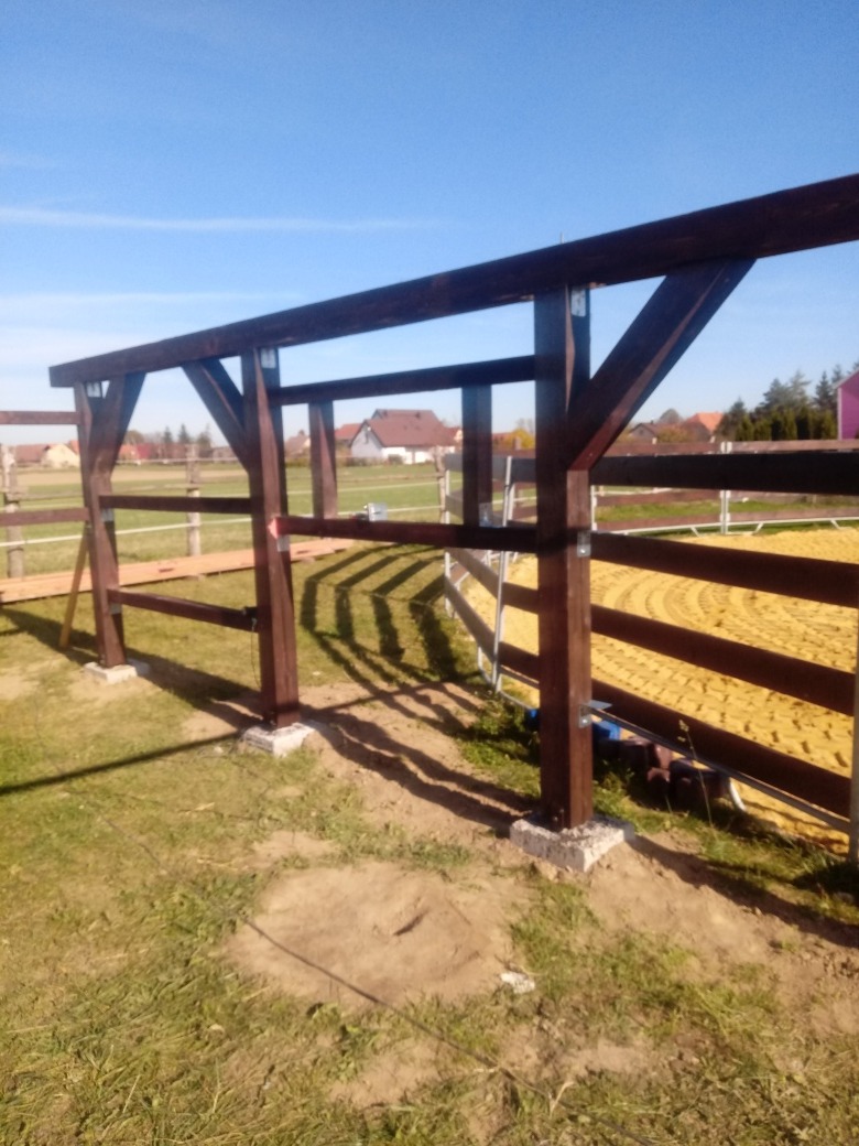 Wiązary ciemnobrązowej drewnianej konstrukcji wiaty/garażu na betonowych podstawach, widok z boku na tle pastwiska i domów w oddali, w słoneczny dzień.
