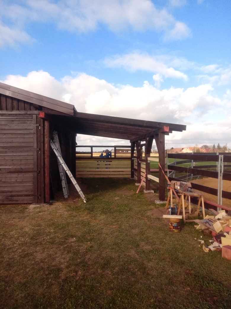 Drewniana wiata/garaż w trakcie budowy, widoczna drabina oparta o ścianę, narzędzia i materiały budowlane na trawie, częściowo zadaszona konstrukcja.
