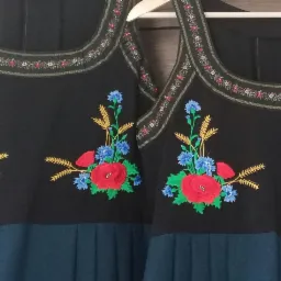 Dwie czarne sukienki z haftowanym bukietem polnych kwiat&oacute;w (mak, chaber, zboże) na wysokości klatki piersiowej i wykończeniem dekoltu drobnym haftem kwiatowym, wiszące na wieszakach.