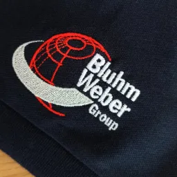 Granatowa koszulka polo z wyhaftowanym logo firmy 'Bluhm Weber Group', przedstawiającym stylizowaną kulę ziemską z czerwoną siatką geograficzną i srebrnym półksiężycem.