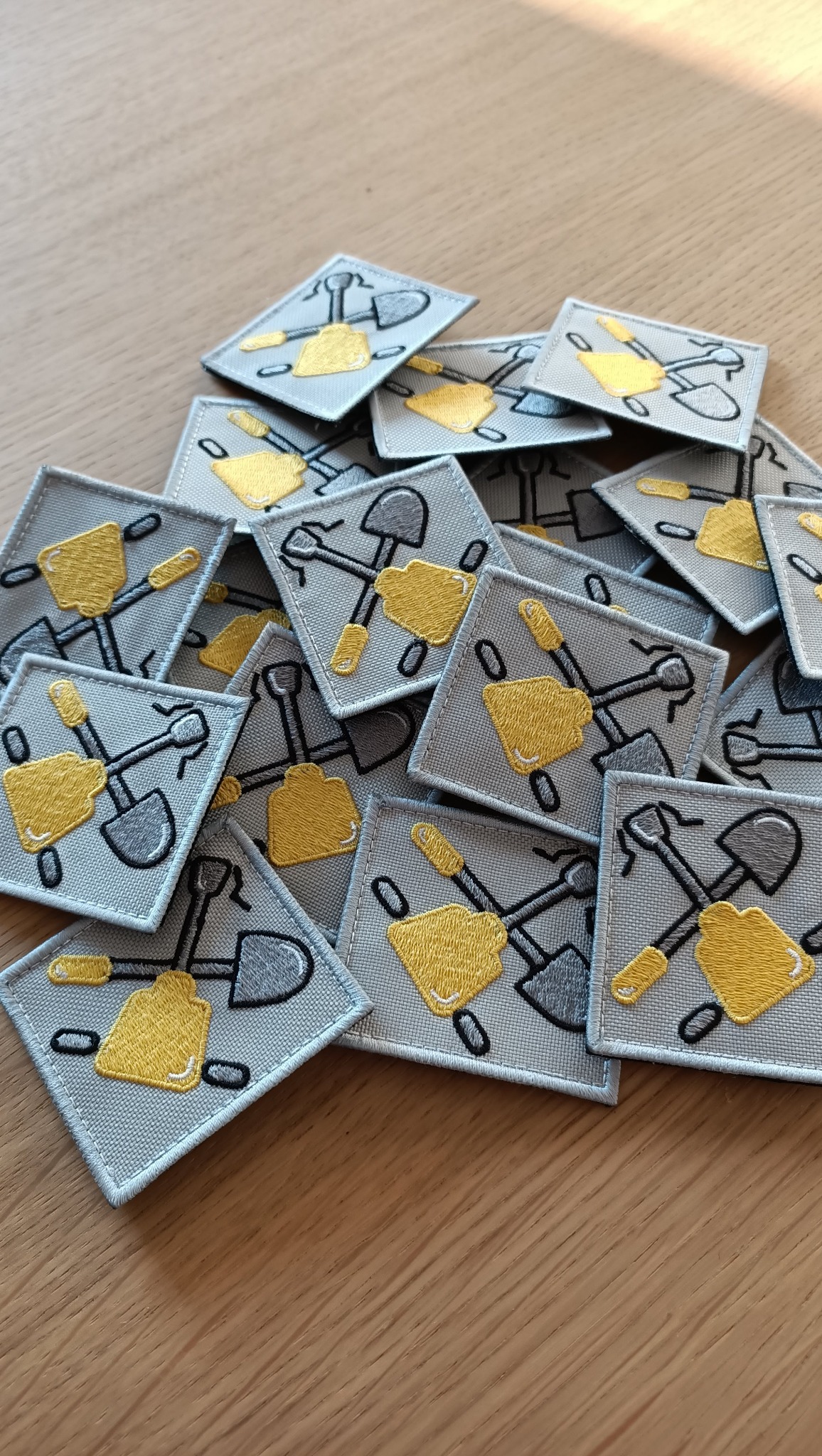 Stos kwadratowych, szarych naszywek z haftowanym motywem skrzyżowanej złotej łopaty i szpadla na drewnianym stole.