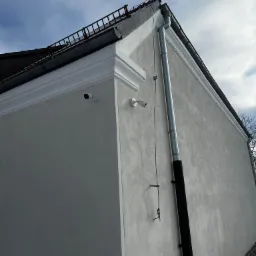 Dwie kamery monitoringu zamontowane na rogu budynku z jasną elewacją, widoczna rynna spustowa i fragment dachu z zabezpieczeniem przeciwśniegowym.