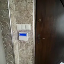 Panel sterowania systemu alarmowego z niebieskim wyświetlaczem, zamontowany obok włączników światła na ścianie wyłożonej kamieniem dekoracyjnym, obok ciemnobrązowych drzwi.