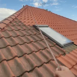 Czerwona dachówka częściowo pokryta brudem i mchem, widoczne okno dachowe i lina trzymana w dłoni, błękitne niebo w tle.