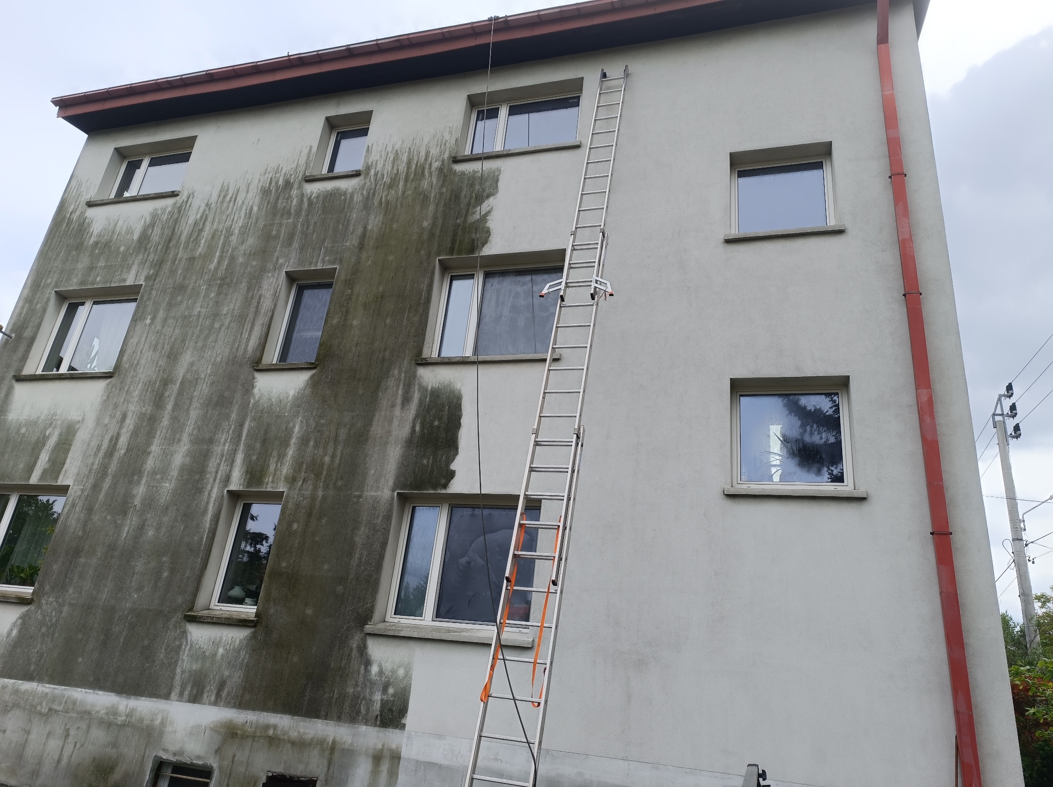 Fragment budynku wielorodzinnego z widocznym efektem czyszczenia elewacji, gdzie lewa strona jest zabrudzona, a prawa czysta, z przystawioną aluminiową drabiną.