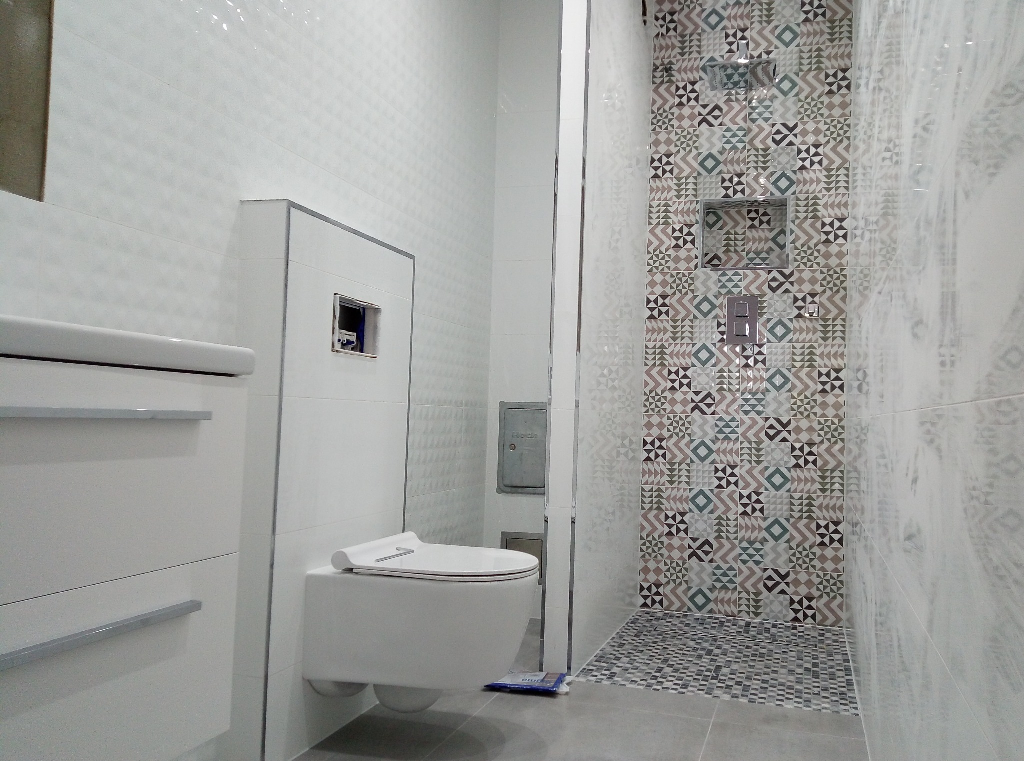 Nowoczesna łazienka z geometrycznymi wzorami na ścianie prysznica, podwieszana toaleta z deską sedesową zintegrowaną z bidetem, białe płytki z delikatnym wzorem na ścianach, szara podłoga i biała...