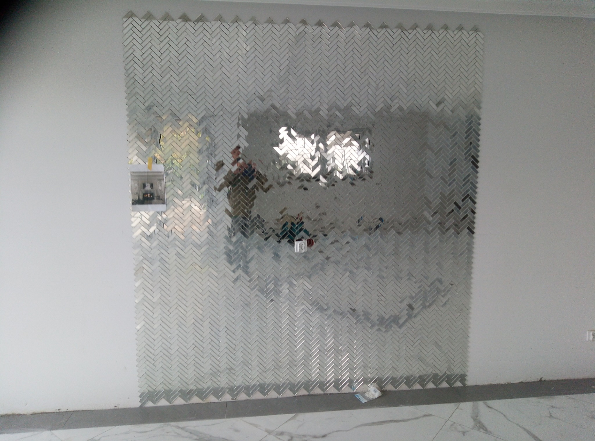 Ściana częściowo pokryta srebrną mozaiką w jodełkę, z widocznym odbiciem wnętrza i wystającym gniazdkiem elektrycznym, na podłodze płytki imitujące marmur.