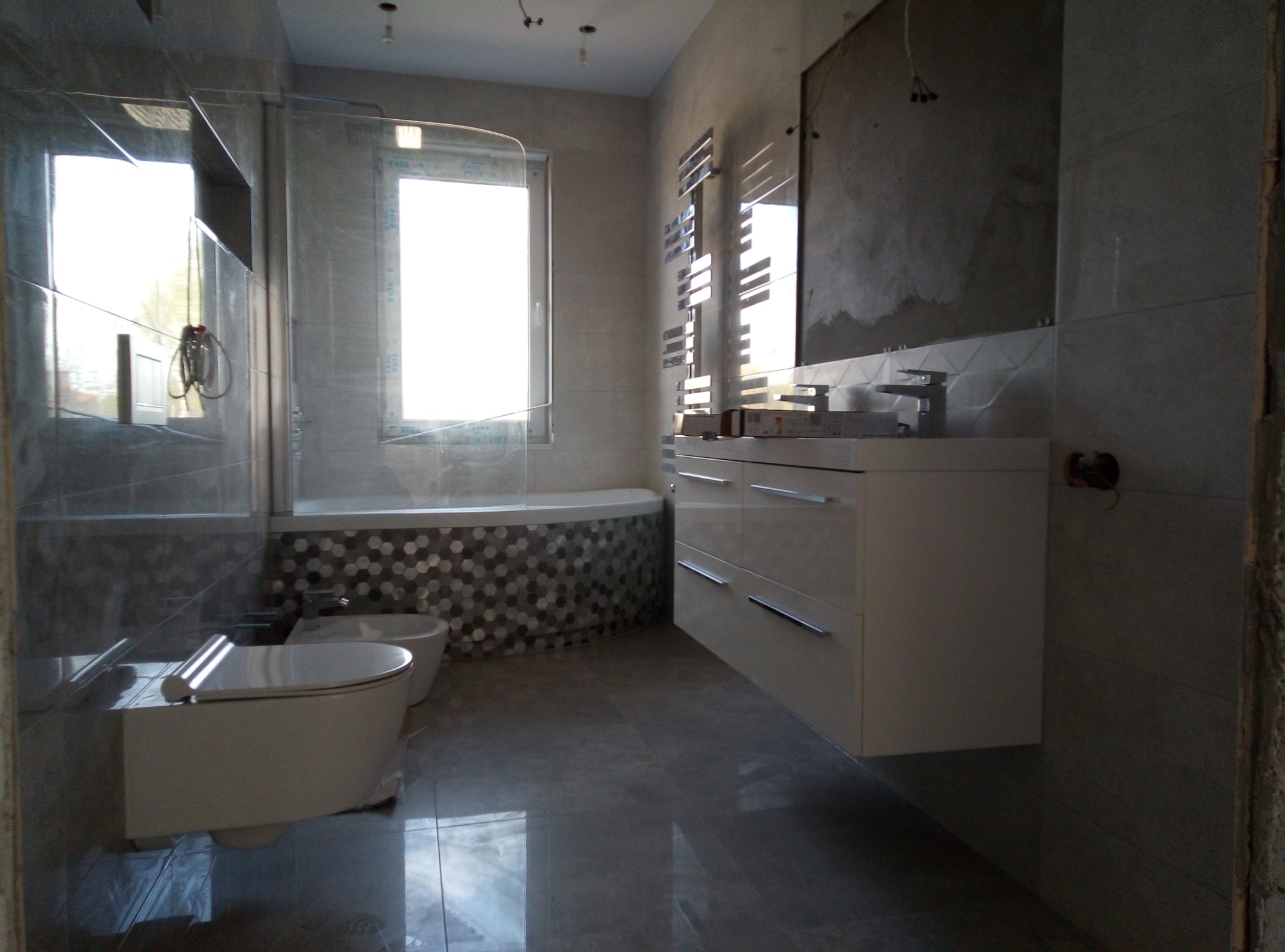 Nowoczesna łazienka w trakcie remontu: szare płytki, biała armatura, mozaika heksagonalna przy wannie, grzejnik drabinkowy, ściana przygotowana pod położenie płytek.