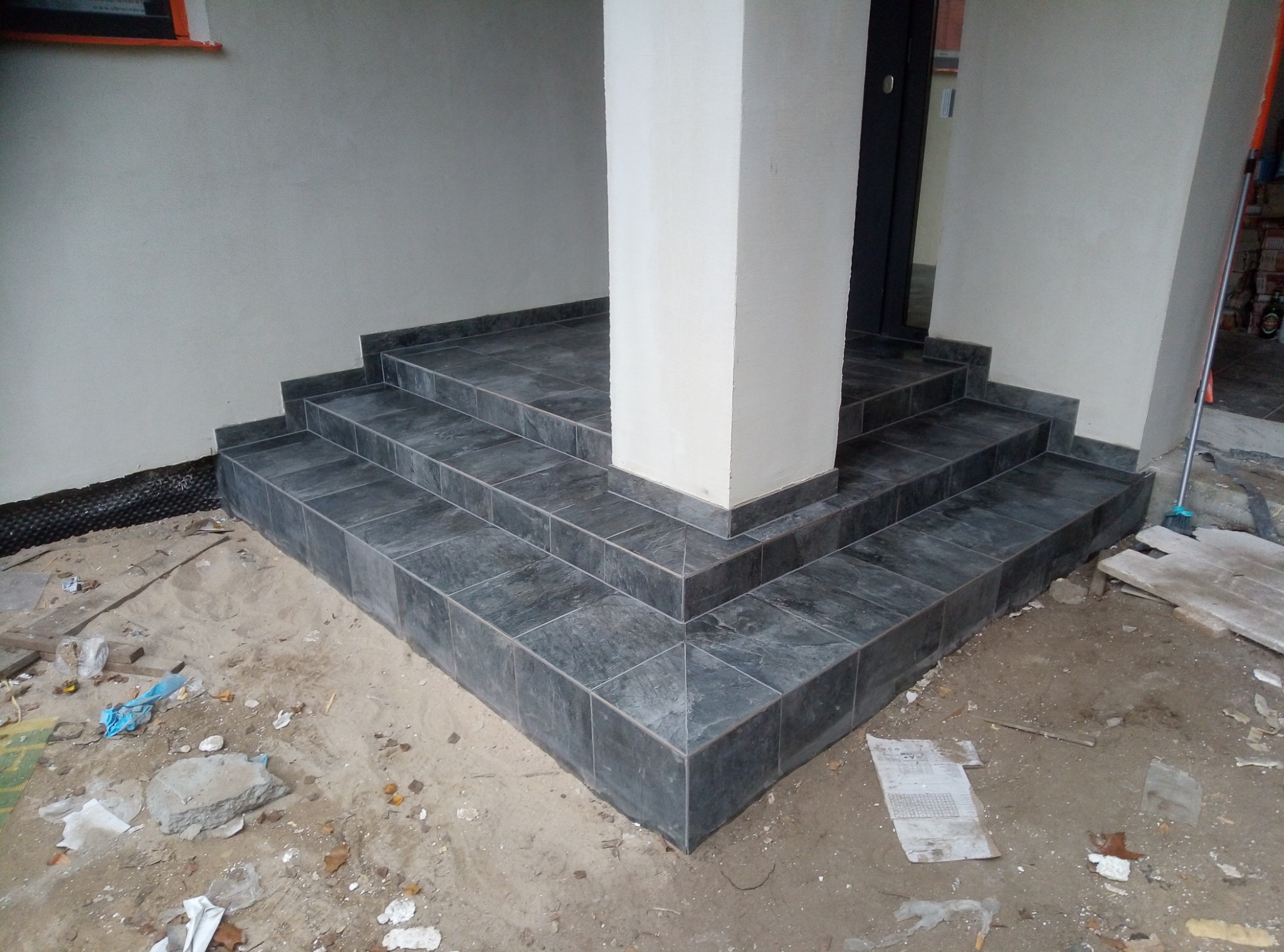 Wykończone schody wejściowe z ciemnoszarych płytek, z kolumną pośrodku, widoczne ślady prac budowlanych na piasku wokół.