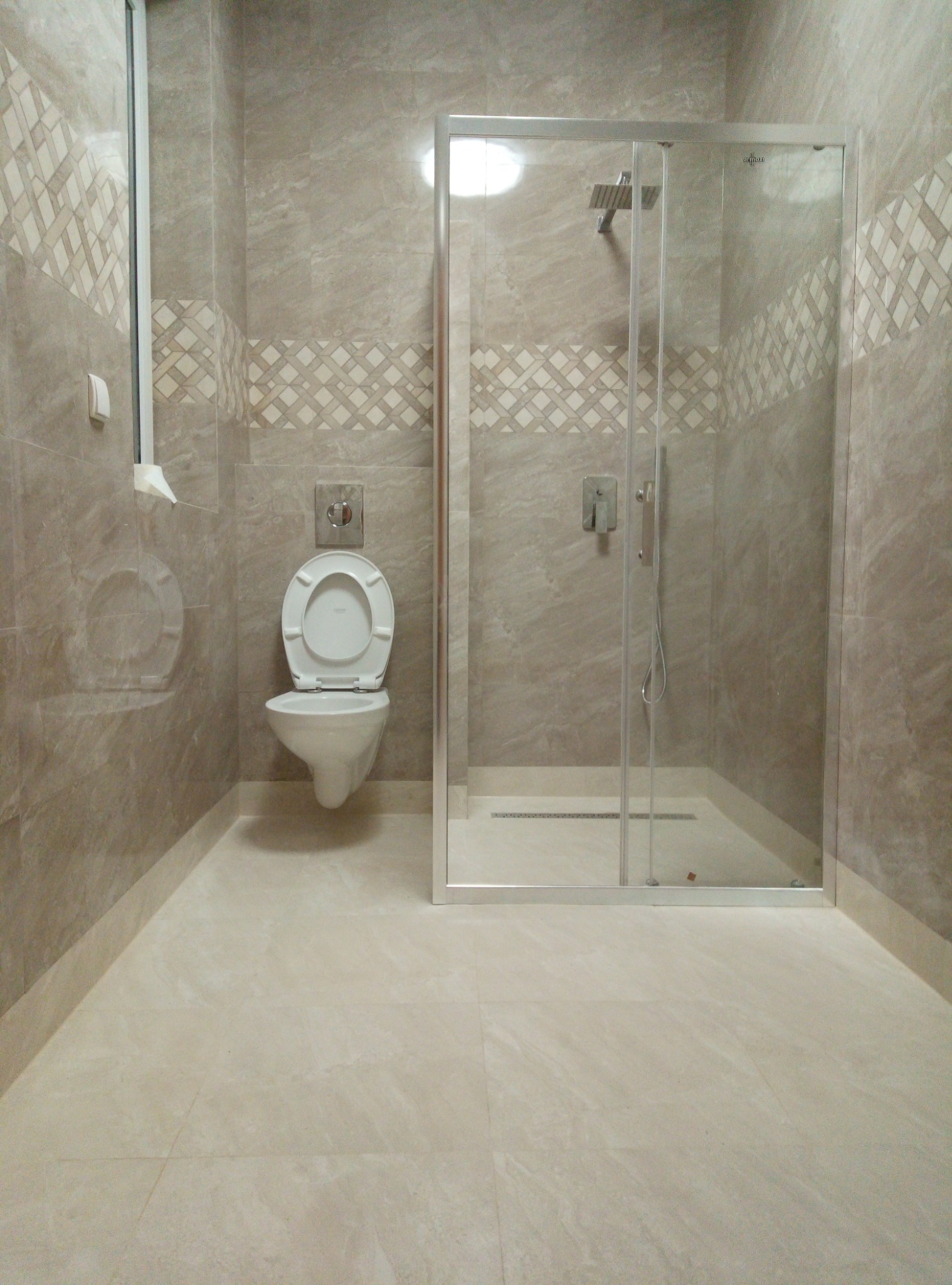 Nowoczesna łazienka wykończona beżowymi płytkami z dekoracyjnym pasem mozaiki, z kabiną prysznicową ze srebrnymi ramami i podwieszaną toaletą z otwartą klapą.