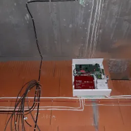 Centrala alarmowa z akumulatorem Zeus ZS-18 zamontowana na ceglanej ścianie w surowym stanie, widoczne kable prowadzone po ścianie i suficie, zabezpieczone pianką montażową w miejscu przejścia...