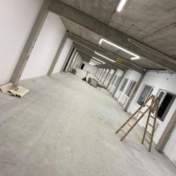 Elektroserwis Arkadiusz Miszczuk - Wnętrze hali przemysłowej w trakcie prac wykończeniowych, widoczne betonowe filary, rzędy okien z grzejnikami, drabina oparta o ścianę oraz lampy jarzeniowe pod sufitem.