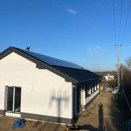 Elektroserwis Arkadiusz Miszczuk - Nowoczesny dom jednorodzinny z panelami słonecznymi na dachu, widoczny z perspektywy ziemi, w tle słup energetyczny i błękitne niebo.