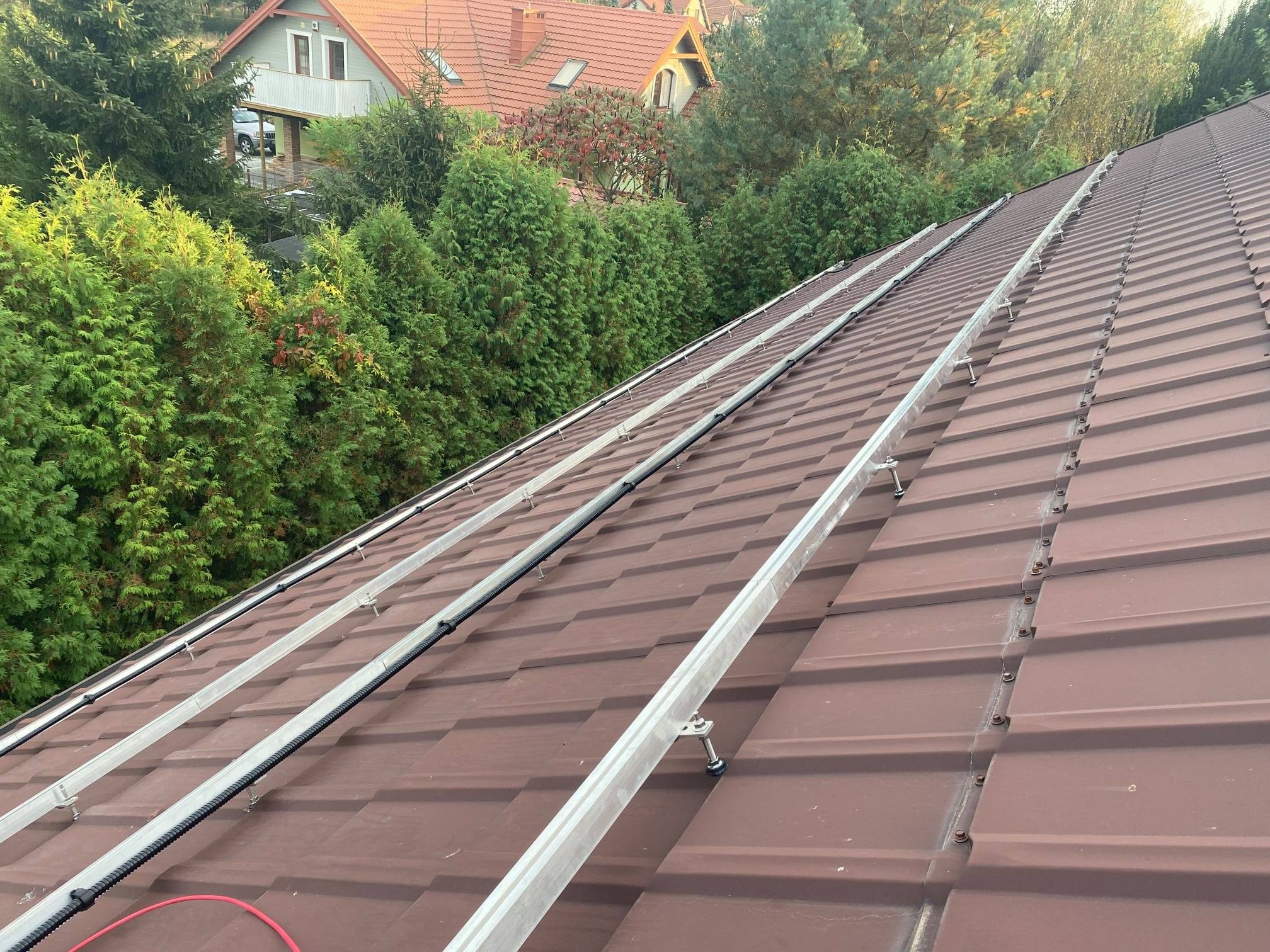 Montaż szyn pod panele słoneczne na brązowym dachu z blachodachówki, widok z góry, w tle zielone tuje i domy z czerwonymi dachami.