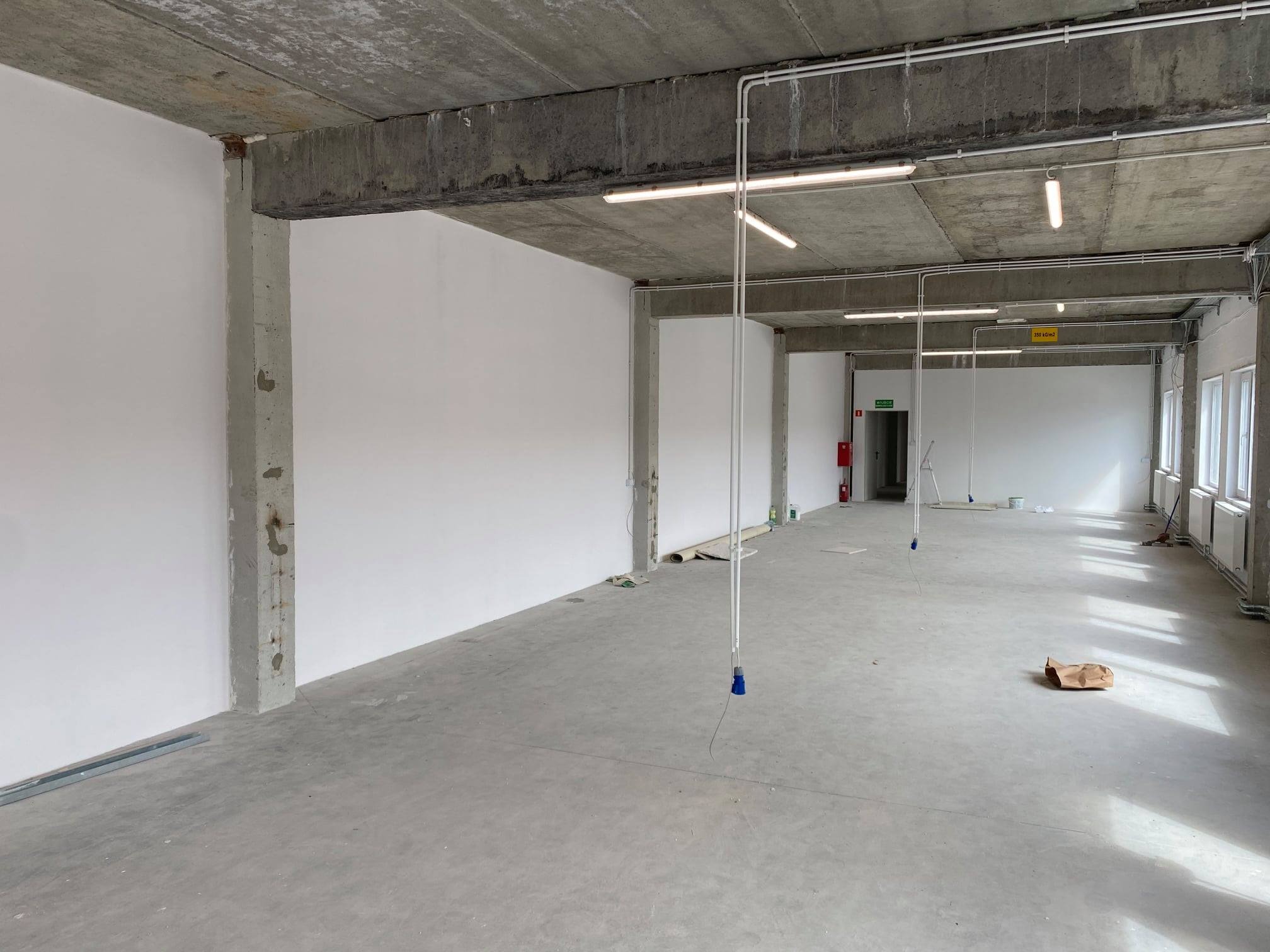 Surowe wnętrze w trakcie prac wykończeniowych: betonowy strop z widocznymi instalacjami elektrycznymi, białe ściany, surowy beton na podłodze, wiszące kable z niebieskimi końcówkami, oraz okna po...