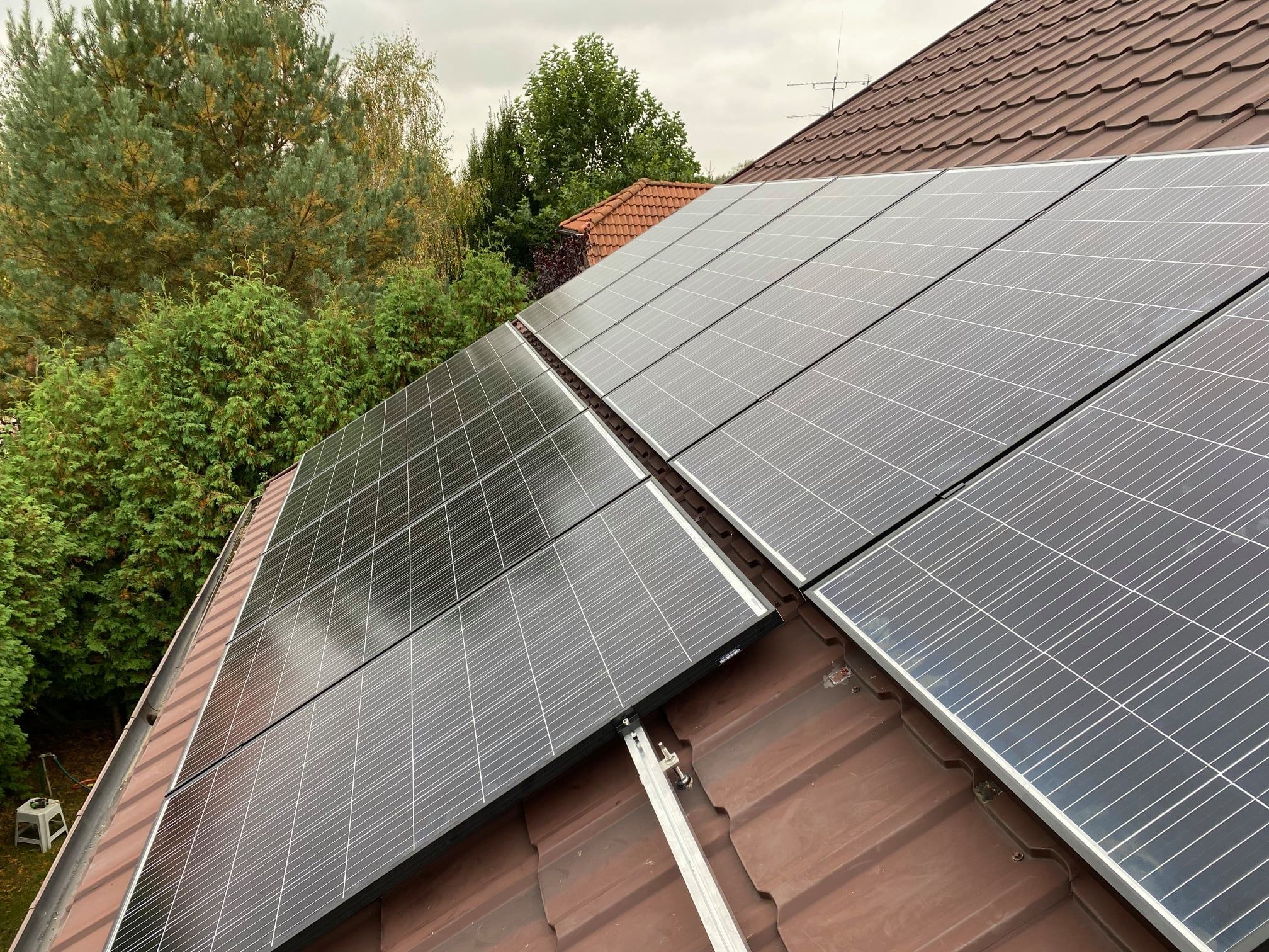 Panele słoneczne zamontowane na brązowym dachu z dachówki ceramicznej, widok z góry, w tle zieleń drzew i fragment dachu sąsiedniego budynku.