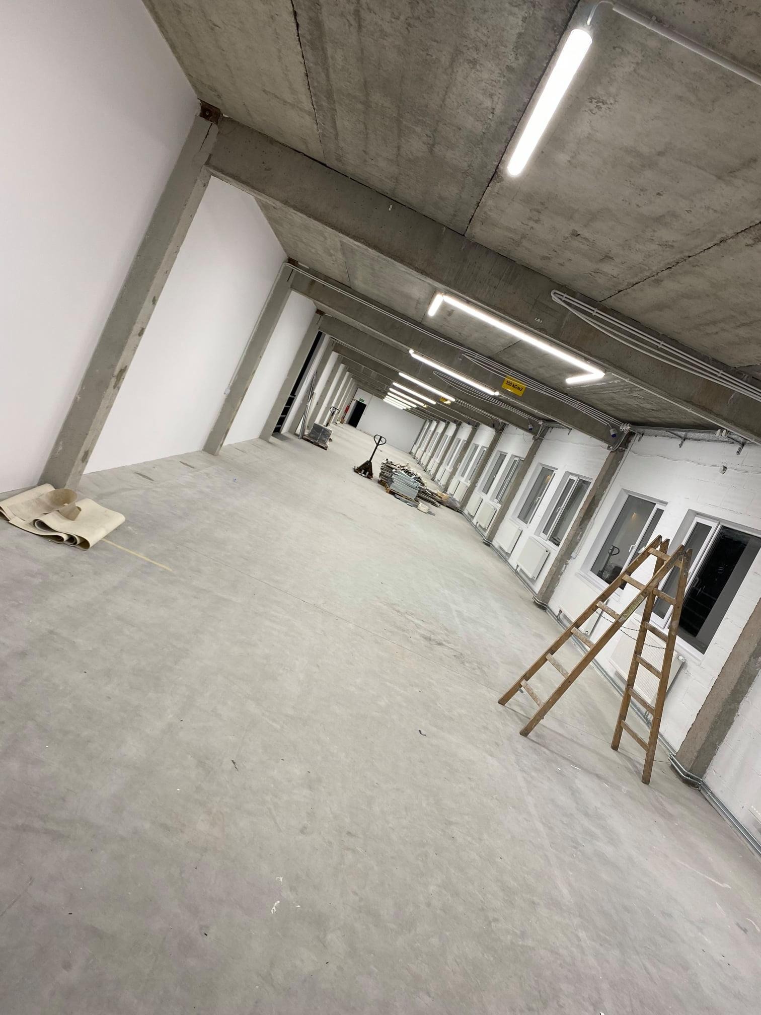 Wnętrze hali przemysłowej w trakcie prac wykończeniowych, widoczne betonowe filary, rzędy okien z grzejnikami, drabina oparta o ścianę oraz lampy jarzeniowe pod sufitem.