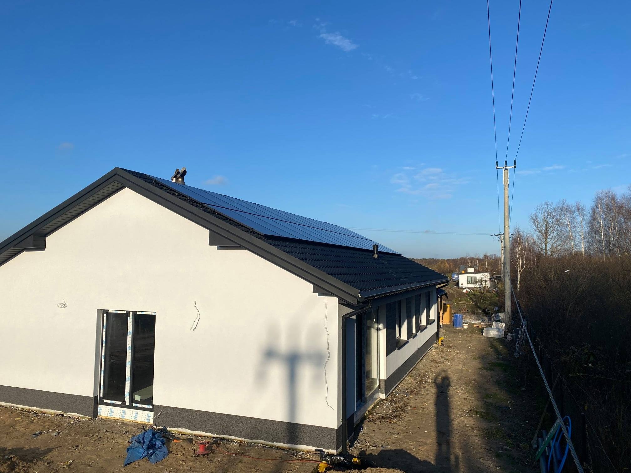 Nowoczesny dom jednorodzinny z panelami słonecznymi na dachu, widoczny z perspektywy ziemi, w tle słup energetyczny i błękitne niebo.