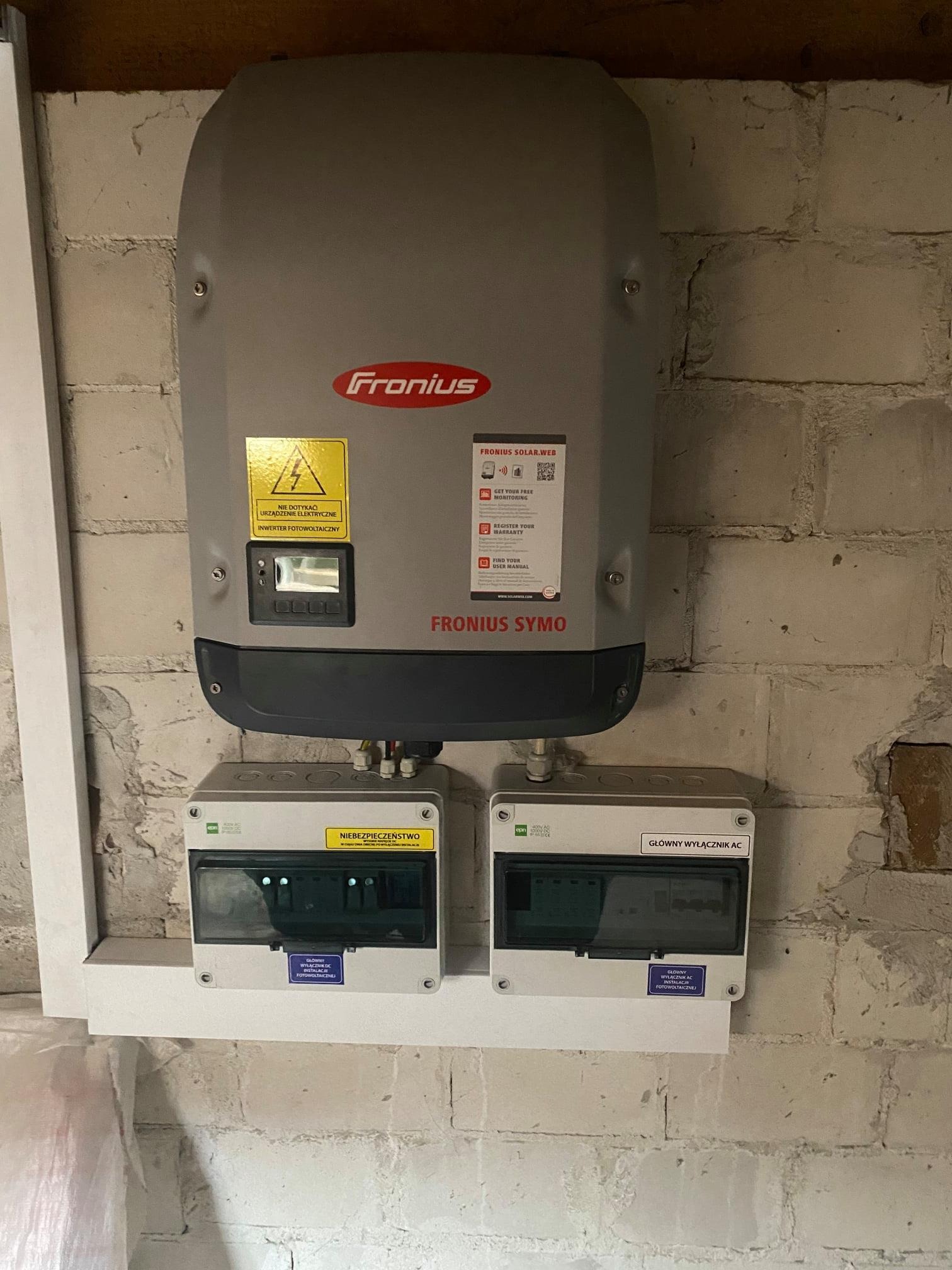 Szary inwerter fotowoltaiczny Fronius Symo zamontowany na białej ceglanej ścianie, z żółtą naklejką ostrzegawczą i dwoma szarymi skrzynkami z napisem 'Główny wyłącznik AC' i 'Wyłącznik DC'.