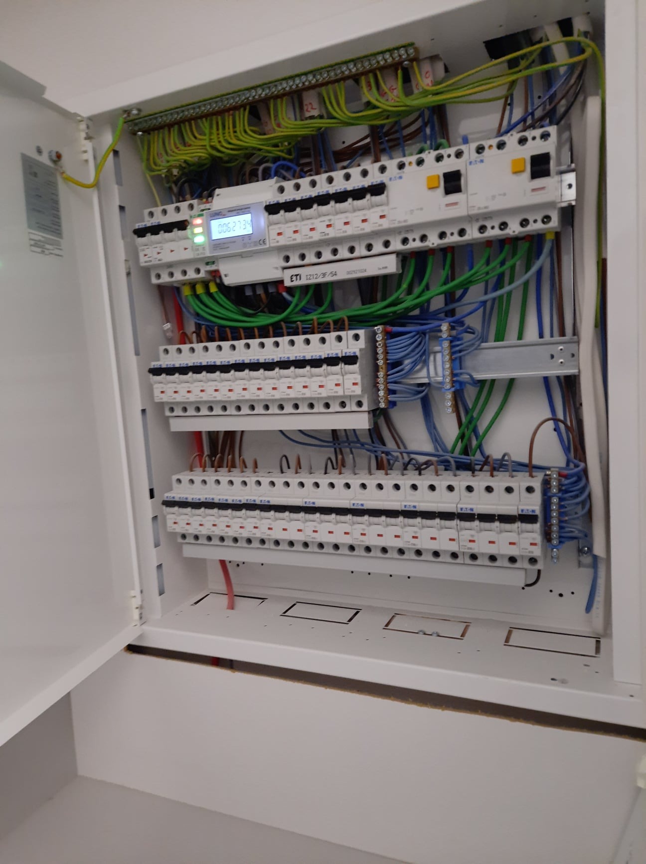 Otwarta skrzynka elektryczna z widocznymi bezpiecznikami, przewodami w kolorach zielonym, niebieskim i brązowym, oraz licznikiem energii z wyświetlaczem wskazującym zużycie.