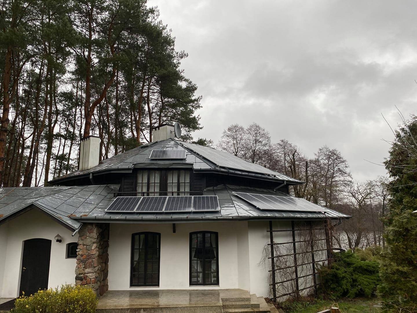 Panele słoneczne zamontowane na dachu dwuspadowym domu z elementami kamiennymi, w tle wysokie drzewa i pochmurne niebo.