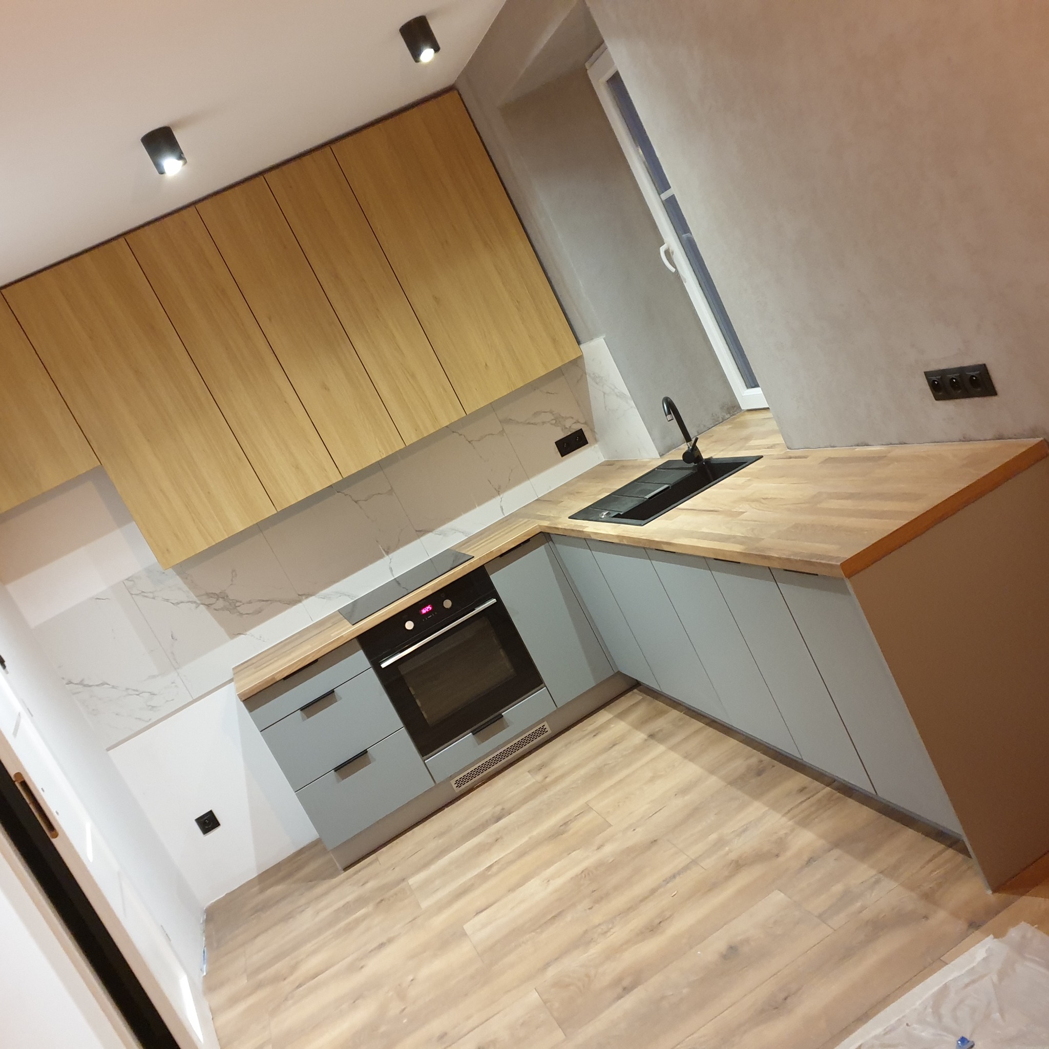 Nowoczesna kuchnia z drewnianymi szafkami górnymi i szarymi dolnymi, jasne drewniane blaty, marmurowe płytki na ścianie, drewniana podłoga, czarna armatura i piekarnik.