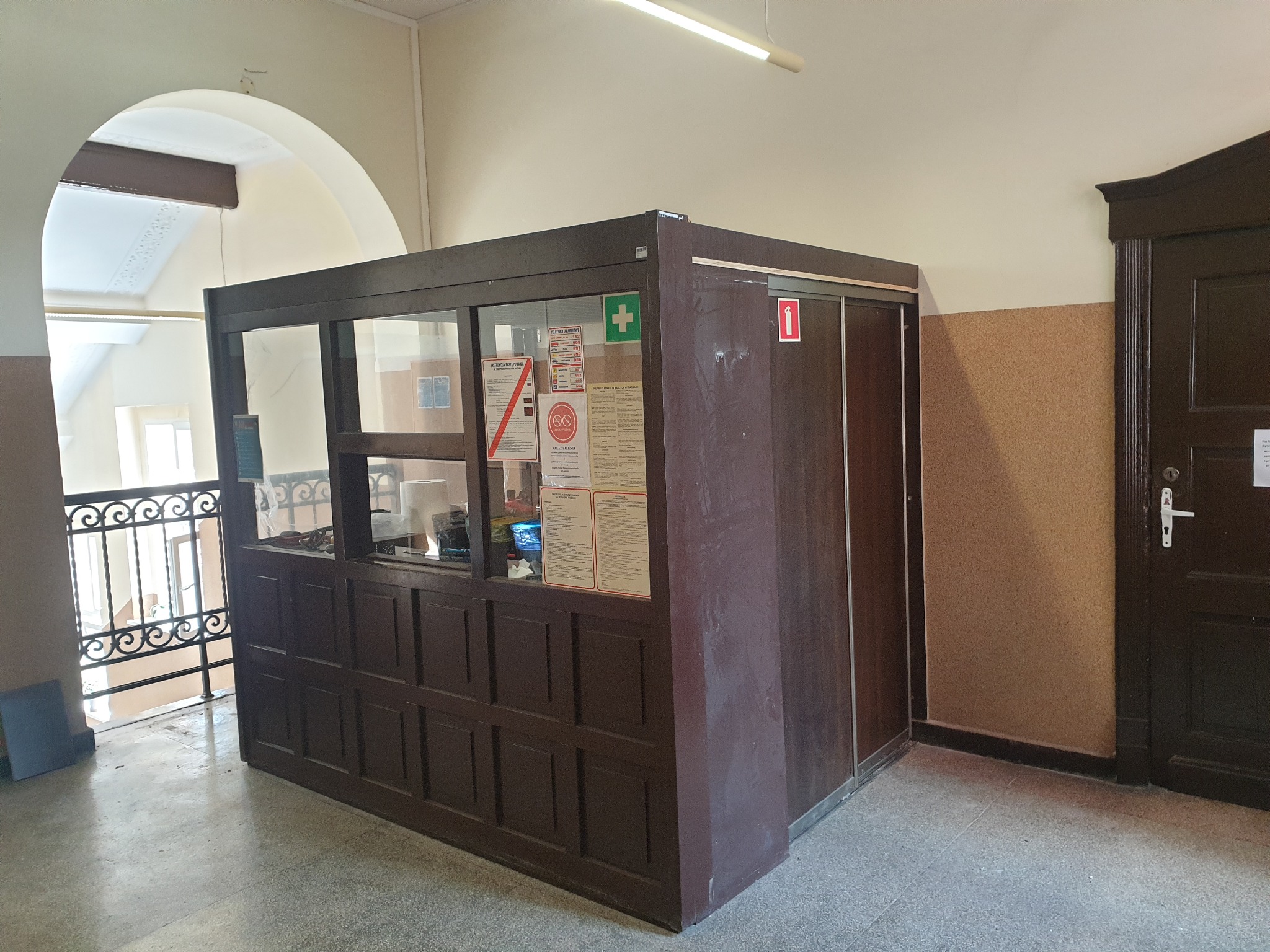 Recepcja z brązowych paneli z szybami, umieszczona na korytarzu w budynku, widoczne dokumenty za szybą i metalowa balustrada w tle.