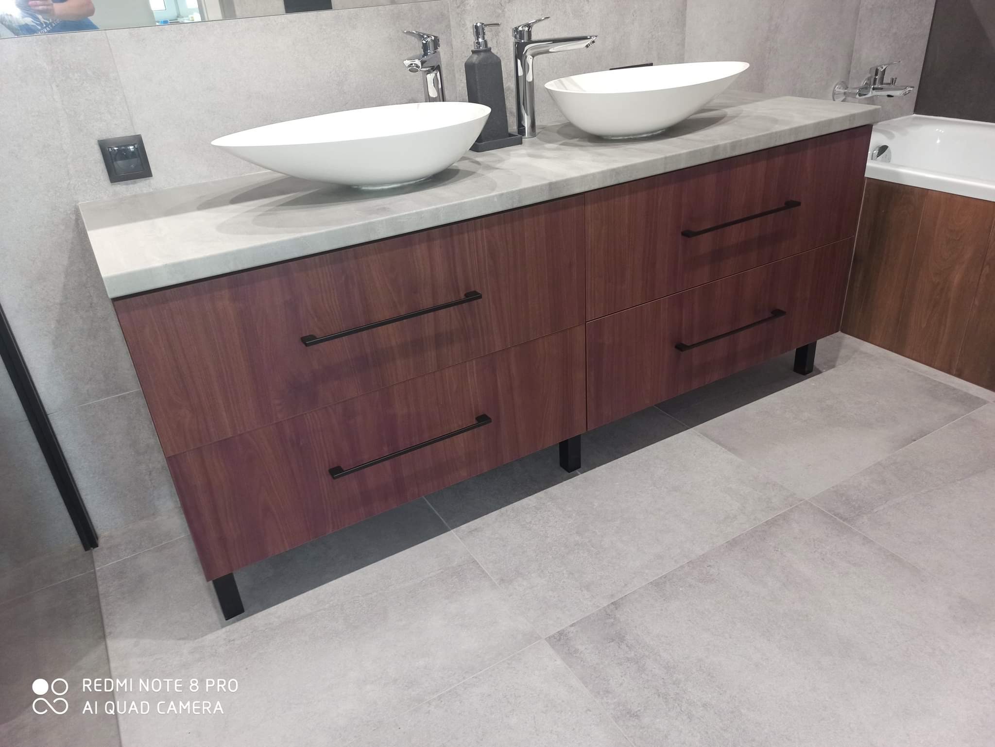 Nowoczesna łazienka z szafką pod umywalką z drewna w odcieniach brązu, szary blat z dwoma białymi umywalkami nablatowymi, minimalistyczne czarne uchwyty i bateria, szare płytki na ścianach...