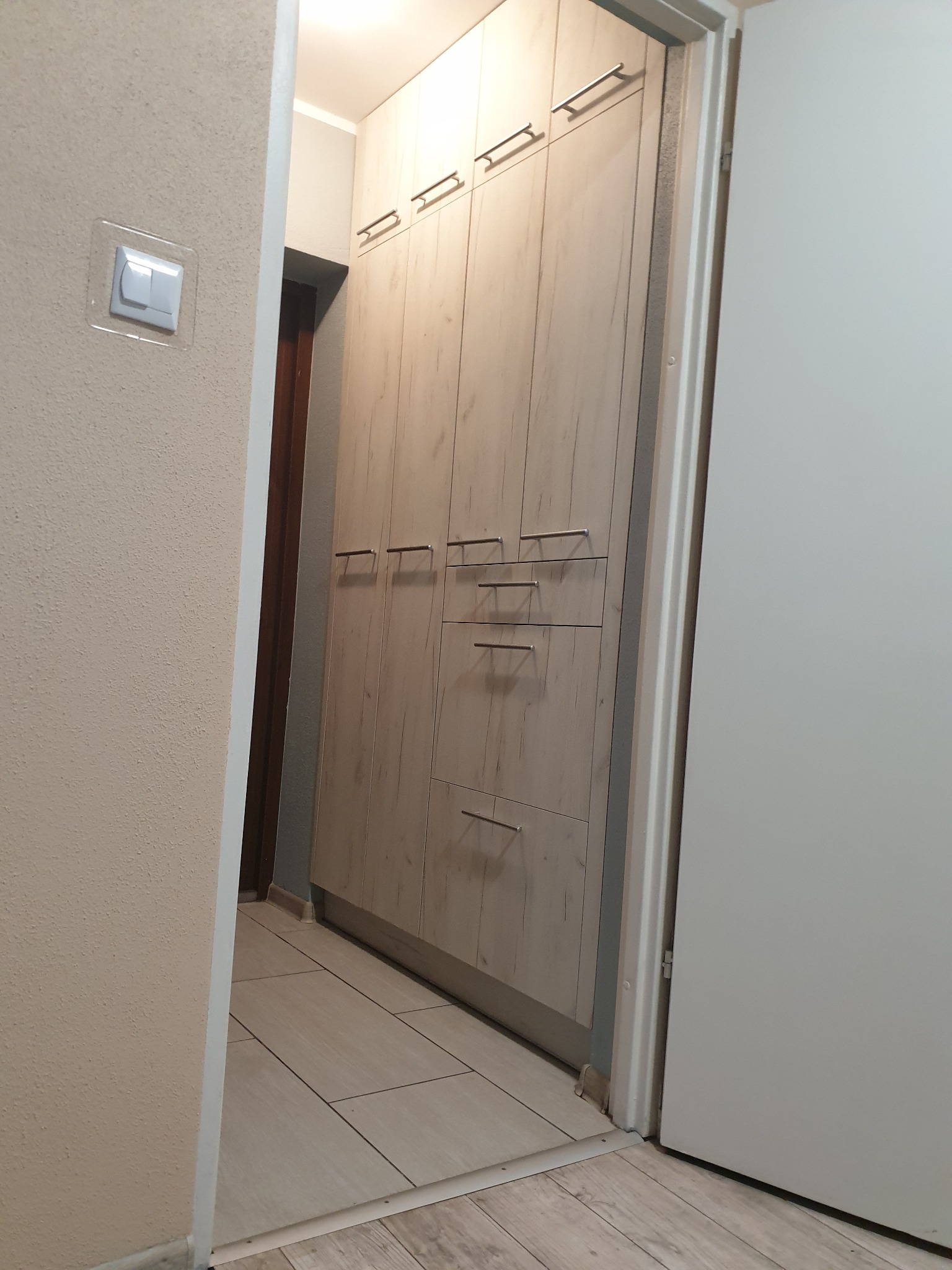 Wysoka szafa na wymiar z jasnego drewna z metalowymi uchwytami, umieszczona w wąskim korytarzu, widoczna z perspektywy wejścia, z włącznikiem światła na ścianie po lewej stronie.