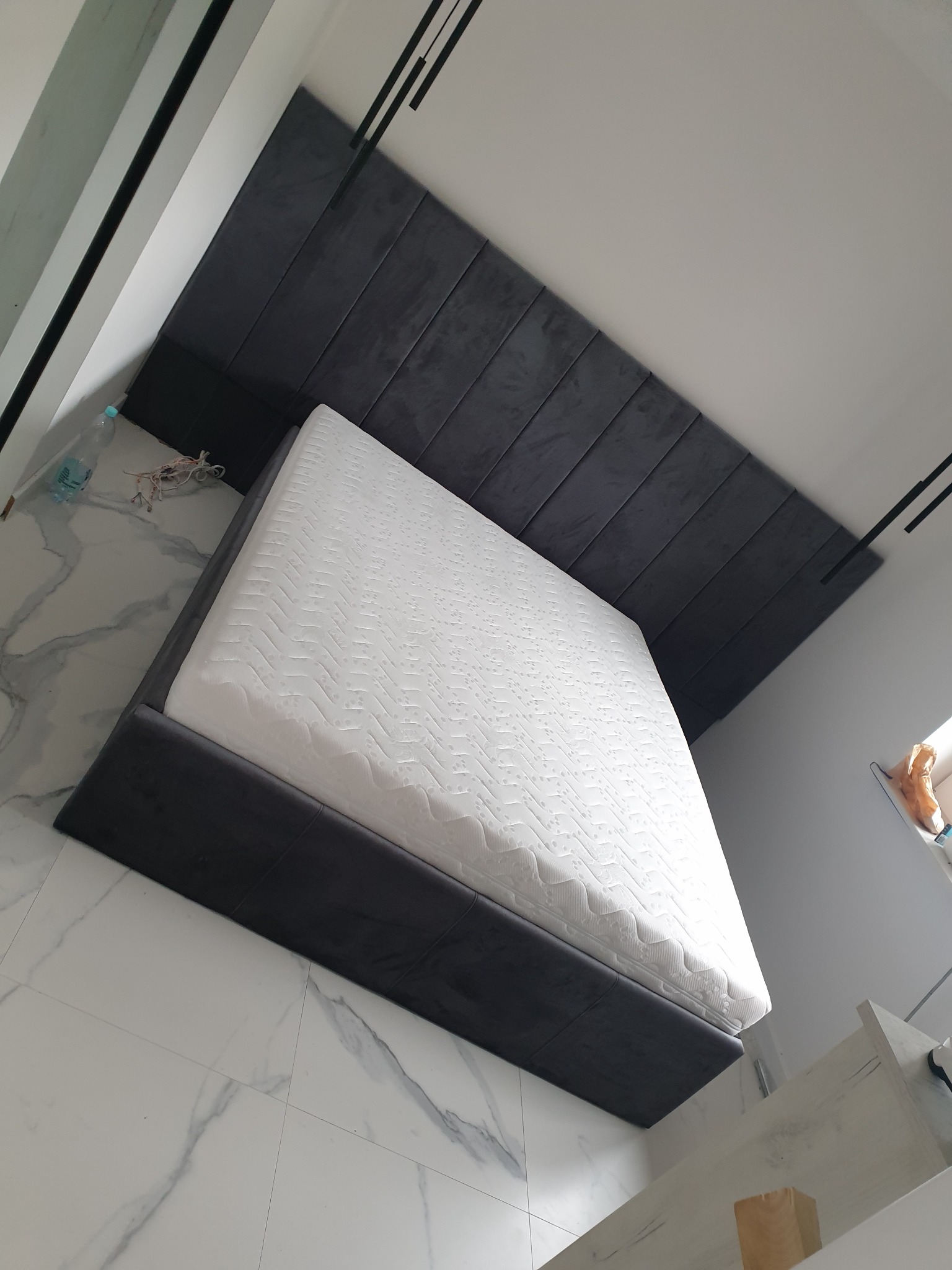 Nowoczesne łóżko z tapicerowanym, ciemnoszarym wezgłowiem, ustawione na marmurowej podłodze, z widocznymi czarnymi lampami sufitowymi.