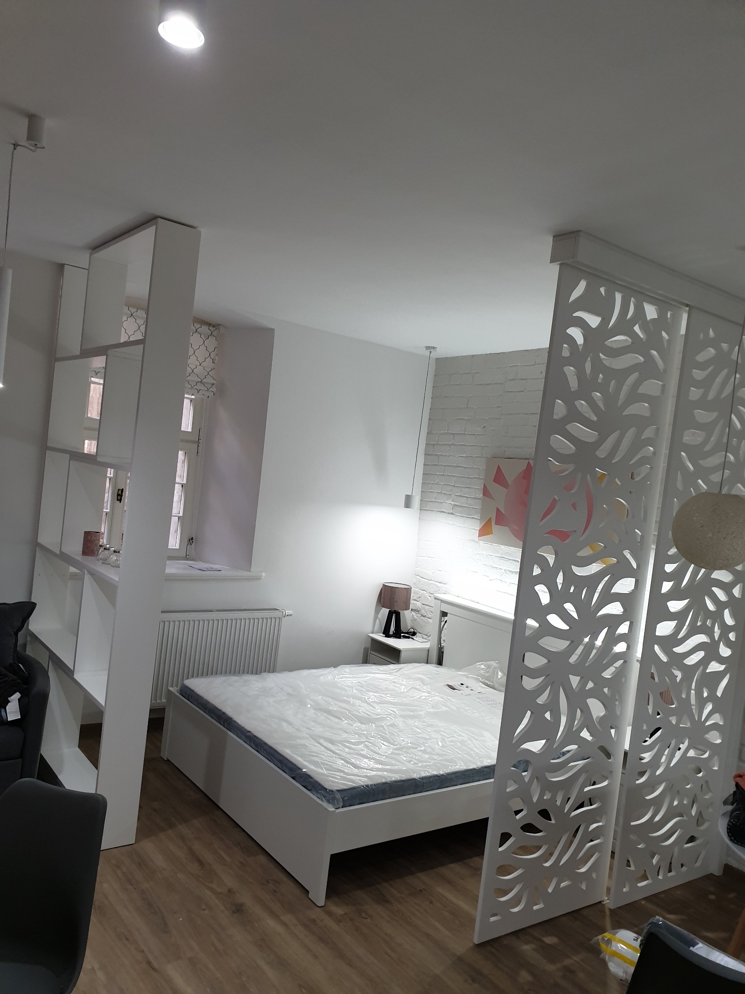 Pokój z białymi meblami oddzielony ażurową ścianką działową, widoczna zabudowa regałowa i łóżko przykryte folią ochronną.