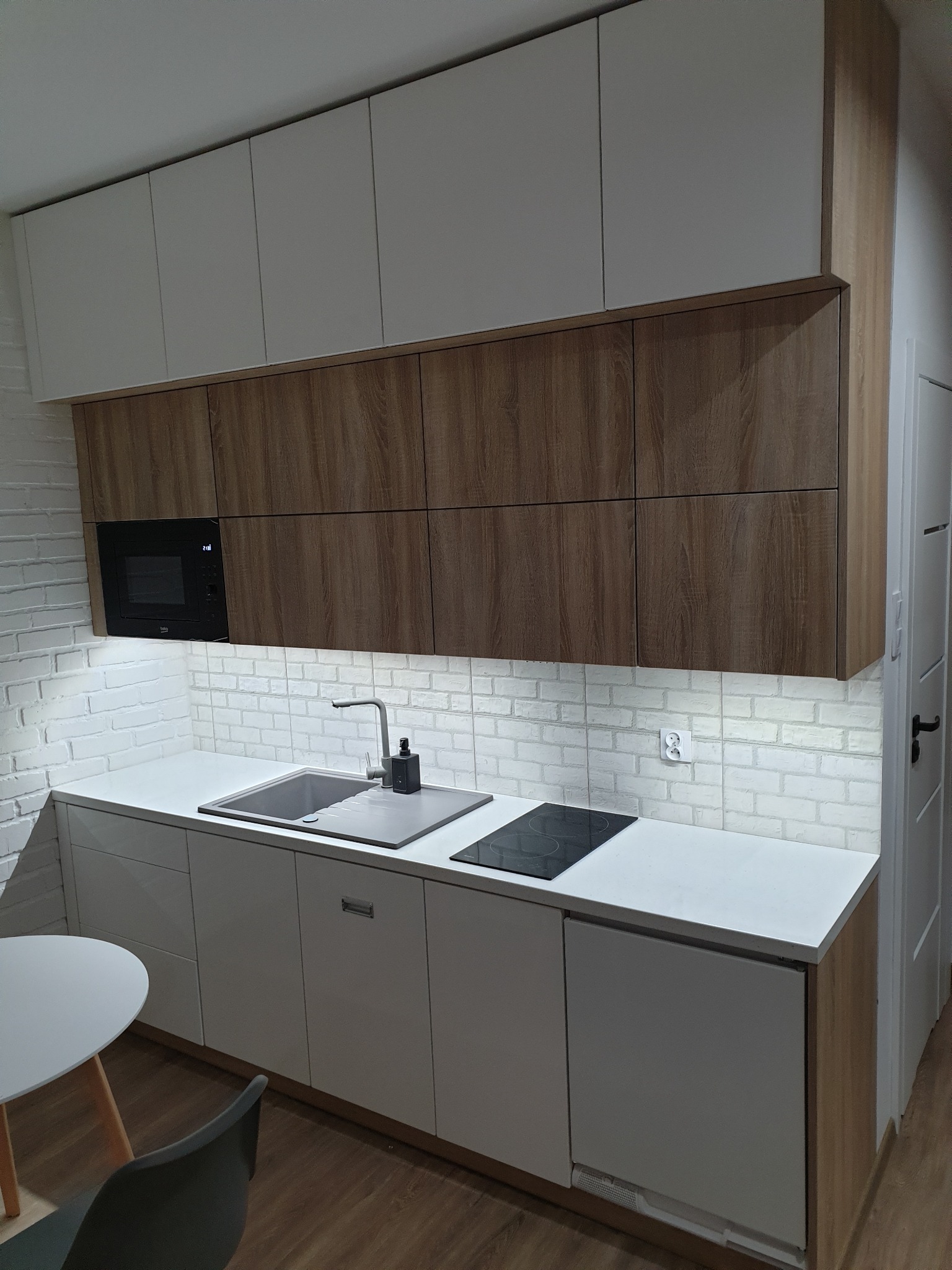 Nowoczesna kuchnia z białymi i drewnianymi szafkami, białą cegłą na ścianie, szarym zlewem i czarną płytą indukcyjną, oświetlona listwą LED pod szafkami wiszącymi.