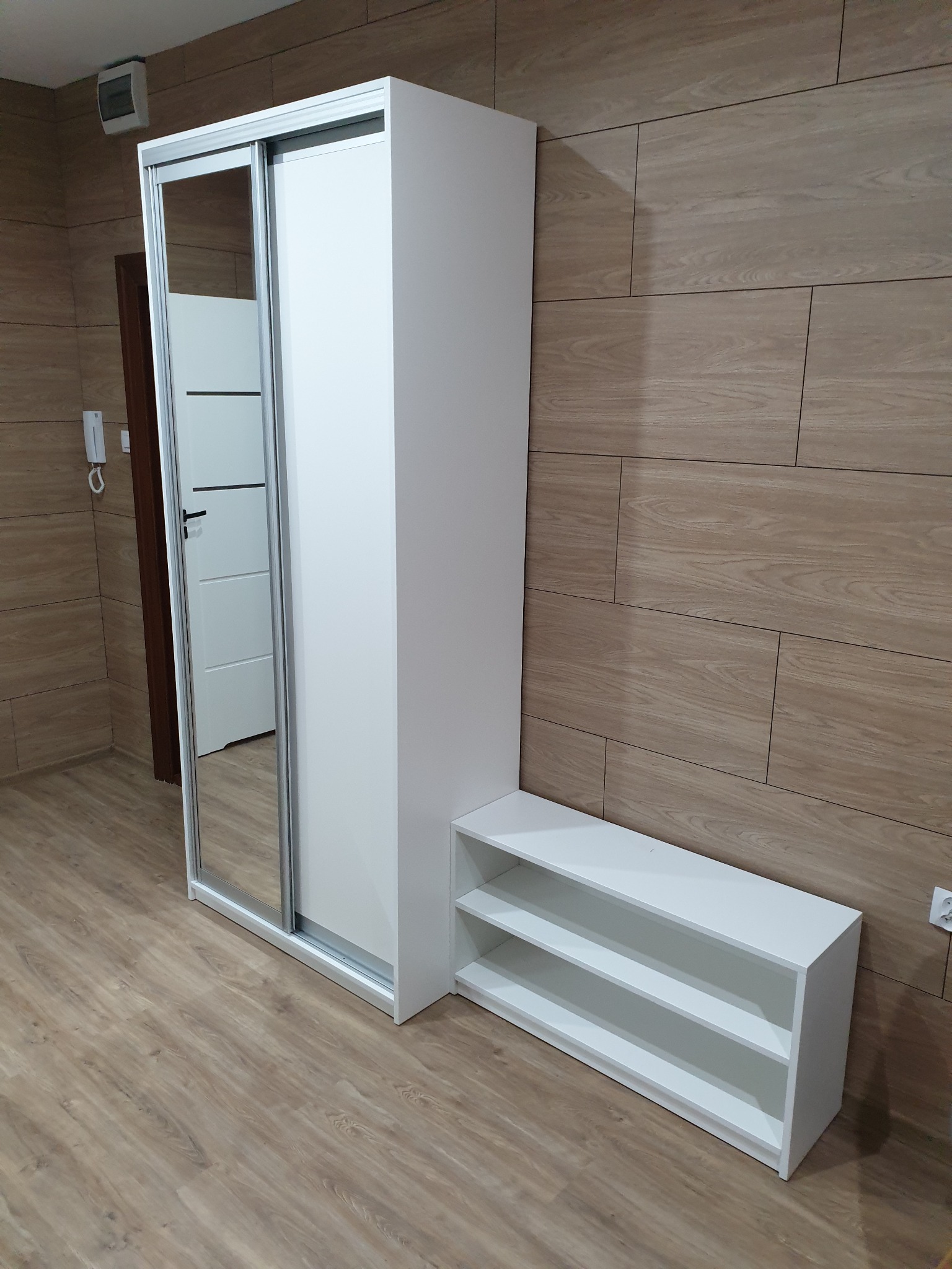 Biała szafa przesuwna z lustrem i regał na buty, ustawiona na drewnopodobnej podłodze przy ścianie z paneli imitujących drewno.